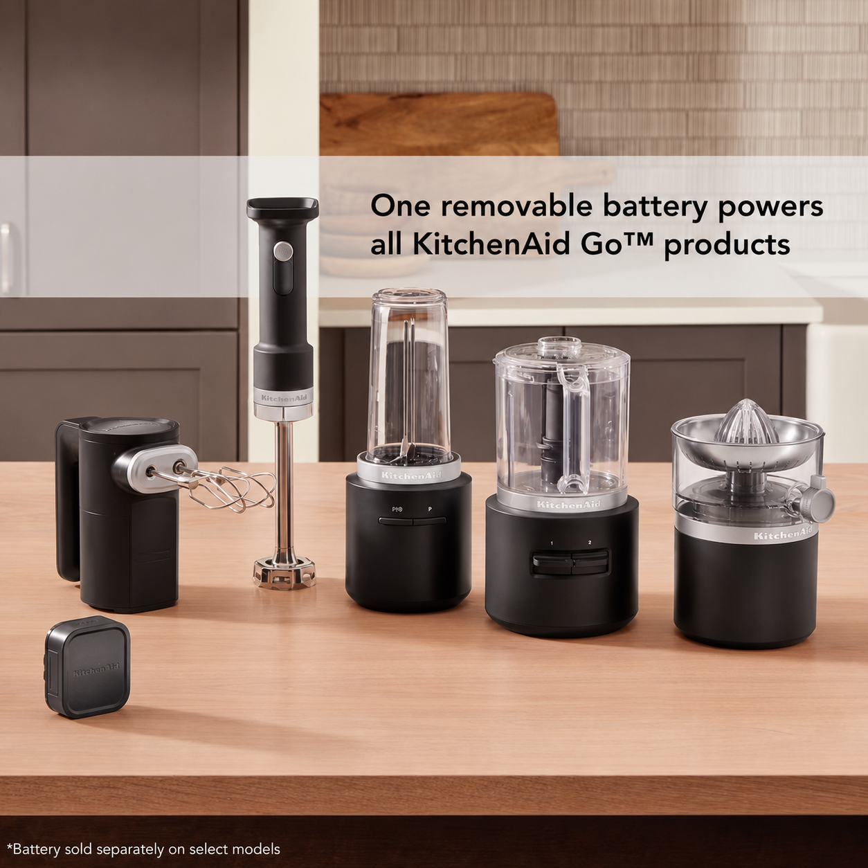 A set of KitchenAid Go products displayed on a wooden countertop in a kitchen setting, including a handheld immersion blender, a blender jar, a food chopper, a citrus juicer, a whisk attachment, and a small square battery. Text on the image reads, 'One removable battery powers all KitchenAid Go products.